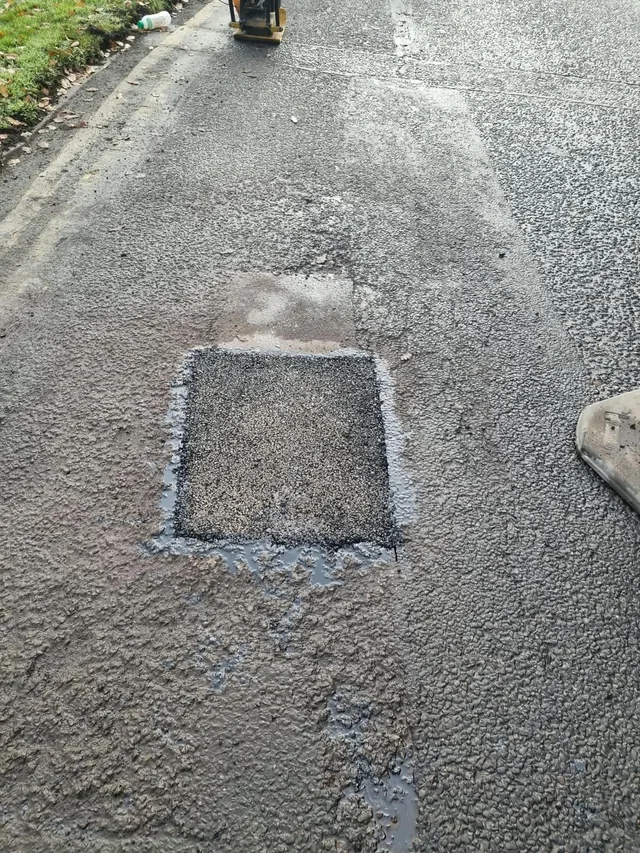 A rectangular asphalt repair patch with a plate compactor nearby